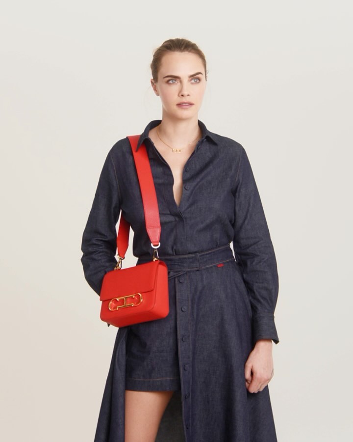 A woman in a dark denim dress stands in a studio with a red cross-body bag, looking off-camera against a neutral background.