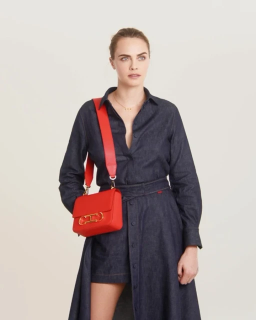 A woman in a dark denim dress stands in a studio with a red cross-body bag, looking off-camera against a neutral background.