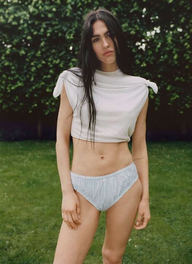 Woman with long dark hair poses outdoors in a light-colored knotted crop top and light blue striped bikini bottoms in natural light.