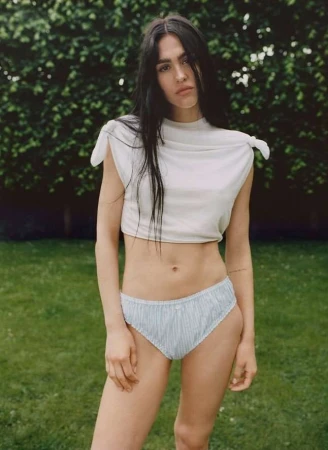 Woman with long dark hair poses outdoors in a light-colored knotted crop top and light blue striped bikini bottoms in natural light.