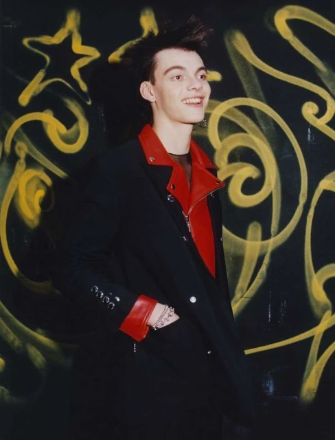 Smiling young adult with distinctive spiky hair and dark jacket with red lapels in front of black wall with yellow graffiti art.