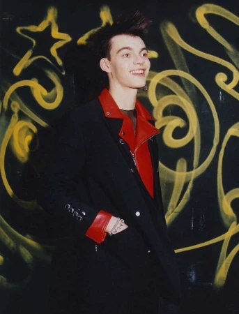 Smiling young adult with distinctive spiky hair and dark jacket with red lapels in front of black wall with yellow graffiti art.