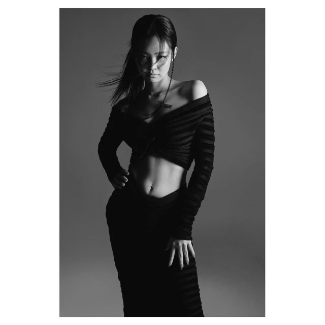 A woman with dark hair poses in a black off-the-shoulder top and skirt against a grey backdrop in a studio portrait, fashion.