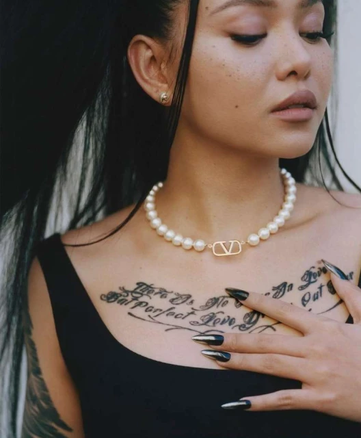 Young woman with an upper chest tattoo poses indoors, wearing pearl jewelry and a black top, against an off-white wall.