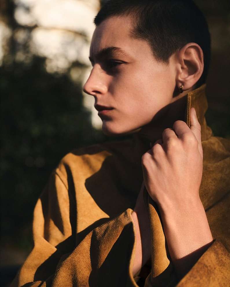 Portrait of a young adult with short dark hair wearing a yellow coat, captured outdoors in a soft, diffused natural light setting.