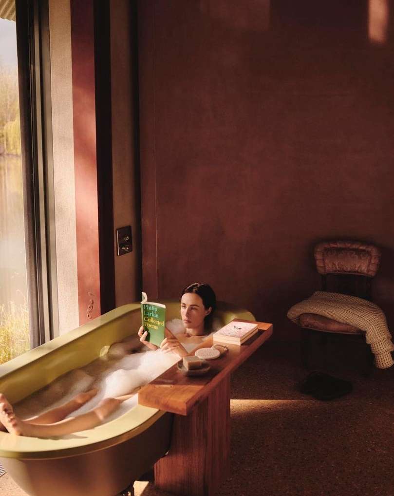 Woman relaxing in a bubble bath, reading a book in a modern bathroom with natural light from a large window overlooking trees.