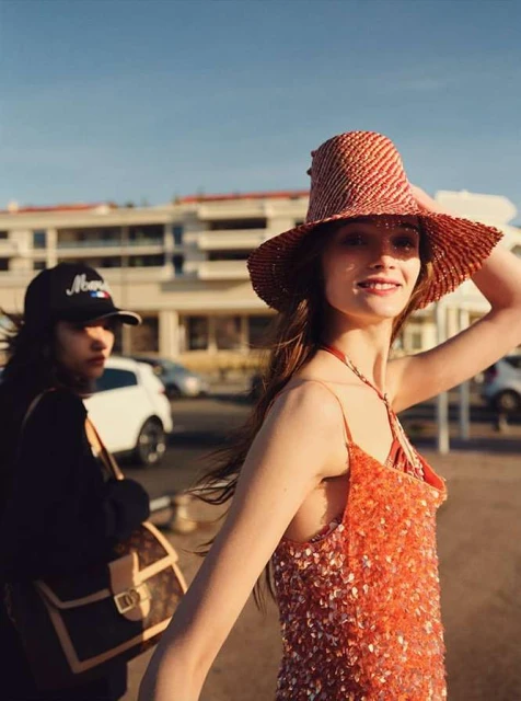 Two women in straw hat and baseball cap stand in sunlight near a building on a clear day, wearing summery orange dress and black jacket.