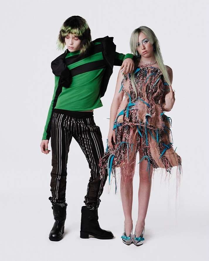 Two fashion models pose in front of a white background. One wears a green striped top and striped pants, the other an avant-garde dress.