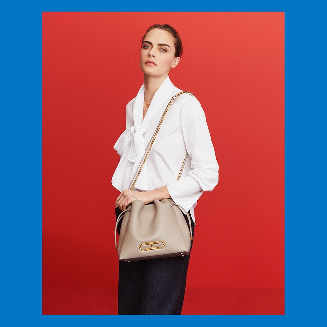 A woman poses against a red backdrop wearing a white blouse, dark jeans, and carrying a light-colored handbag with a gold chain strap.