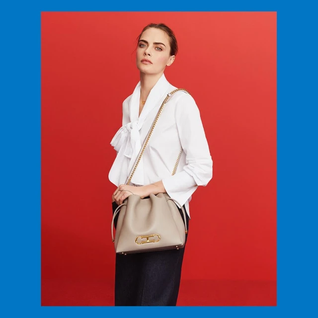 A woman poses against a red backdrop wearing a white blouse, dark jeans, and carrying a light-colored handbag with a gold chain strap.