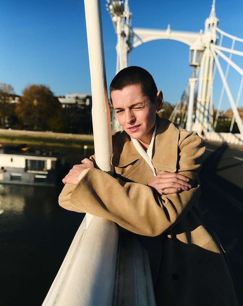 Woman with short hair wearing a beige coat on a bridge with a white structure, blue sky, and river background, posing with arms crossed.