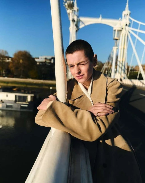 Woman with short hair wearing a beige coat on a bridge with a white structure, blue sky, and river background, posing with arms crossed.