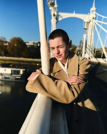 Woman with short hair wearing a beige coat on a bridge with a white structure, blue sky, and river background, posing with arms crossed.