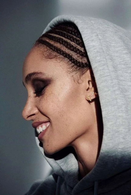 Close-up side profile of a smiling adult woman with braided hair wearing a gray hooded sweatshirt in front of a neutral background.