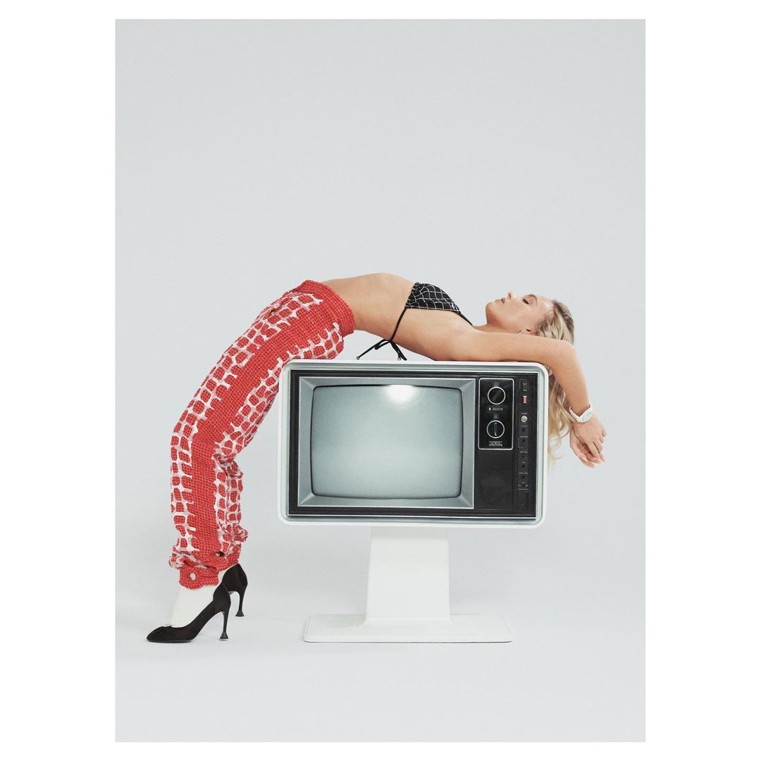 Woman in black bikini top and patterned pants lies in a backbend on a retro television against a bright white backdrop in studio lighting.