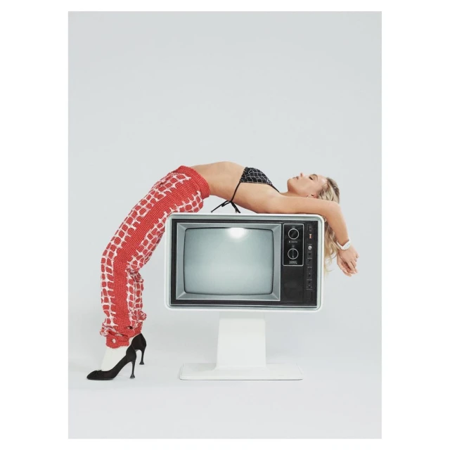 Woman in black bikini top and patterned pants lies in a backbend on a retro television against a bright white backdrop in studio lighting.