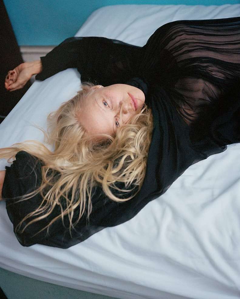 Woman with long blonde hair in a black top reclining on a white bed. Soft lighting emphasizes her relaxed pose and the indoor setting.