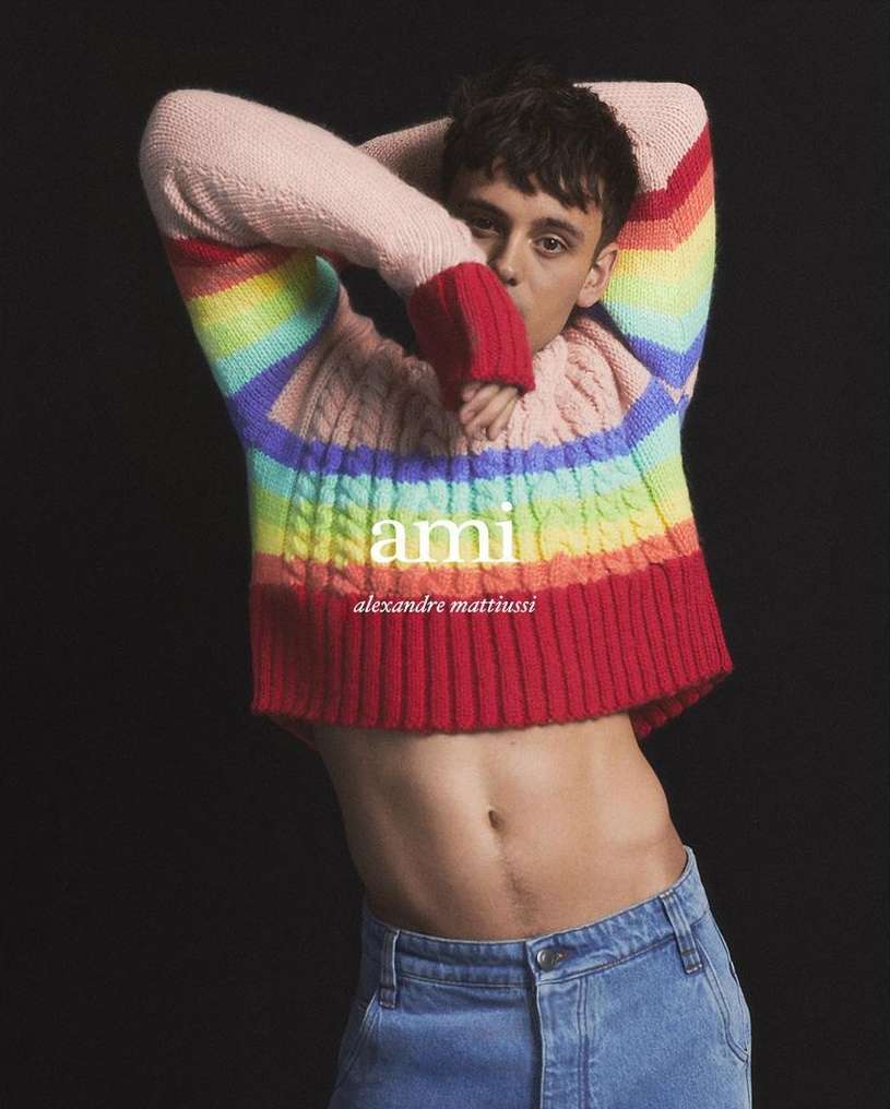 Young man wearing a colorful striped sweater and blue jeans posing against a black background with hands behind head