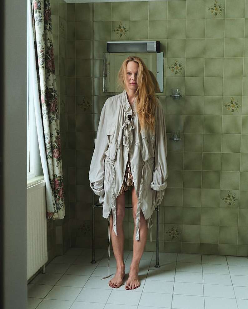 A woman in a jacket stands in a bathroom with green tile, looking directly at the camera with a neutral expression and red hair.