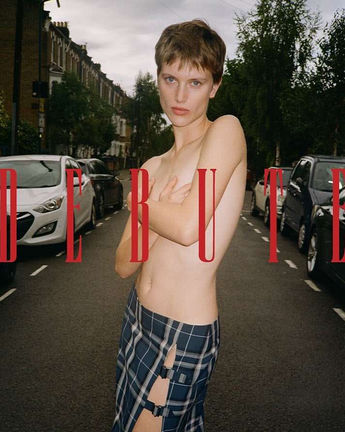 A topless person with short hair wearing a skirt poses on a street, flanked by parked cars, with the word 'DEBUT' superimposed in red.