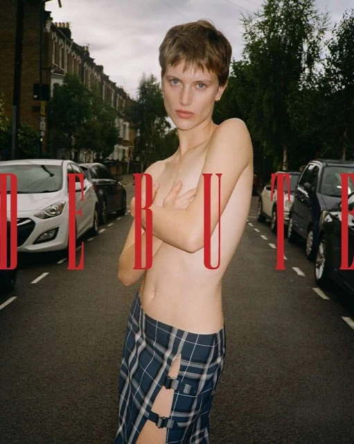 A topless person with short hair wearing a skirt poses on a street, flanked by parked cars, with the word 'DEBUT' superimposed in red.