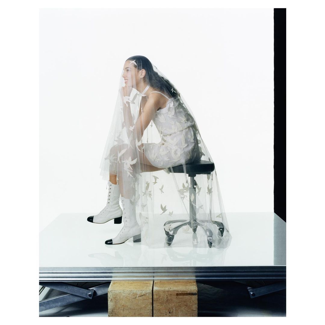 Woman in white lace dress and matching boots sitting on a stool, wearing a sheer patterned veil in a bright photo studio.