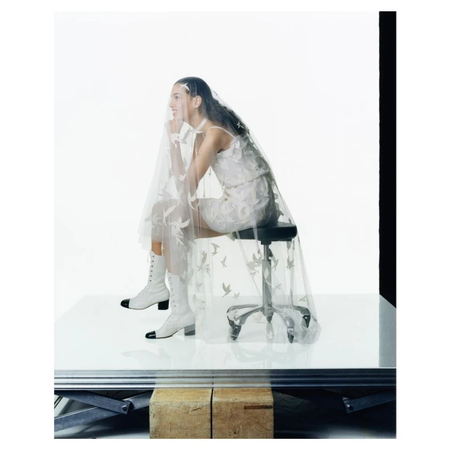 Woman in white lace dress and matching boots sitting on a stool, wearing a sheer patterned veil in a bright photo studio.