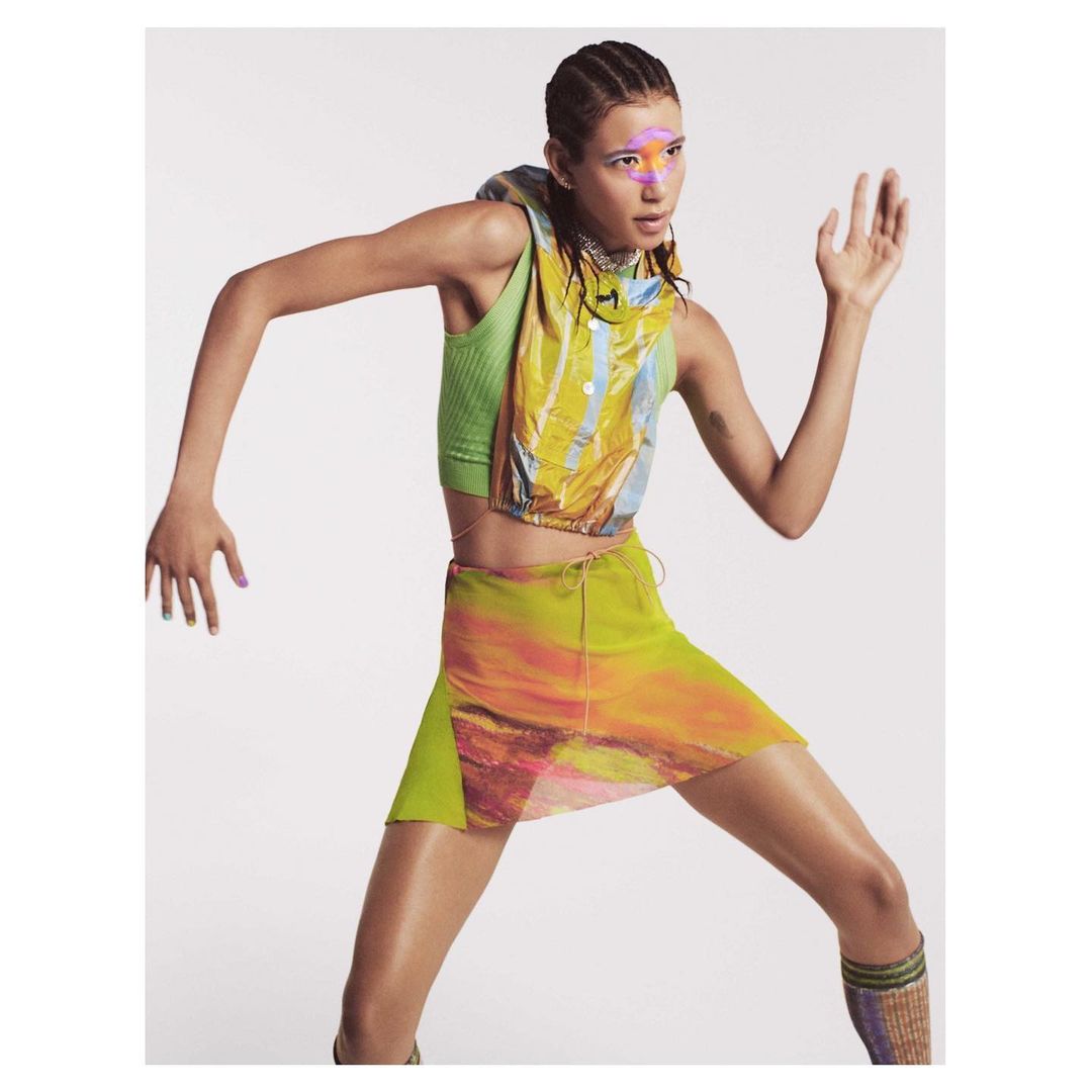 A slender fashion model with face paint poses in a multicolored vest and skirt against a neutral background in a full studio shot.