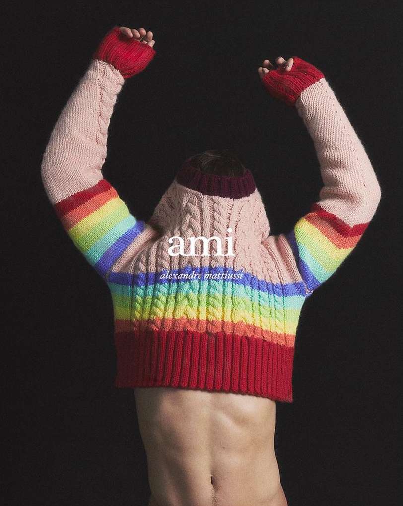Torso of a non-binary person wearing an AMI hooded sweater, arms raised, back to the camera against a dark backdrop. Fashionable and stylish.