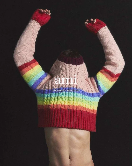 Torso of a non-binary person wearing an AMI hooded sweater, arms raised, back to the camera against a dark backdrop. Fashionable and stylish.