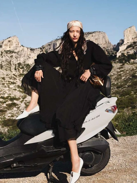 An adult woman in a black dress and headscarf sits on a gray scooter with a backdrop of rock formations under a blue sky on a sunny day.