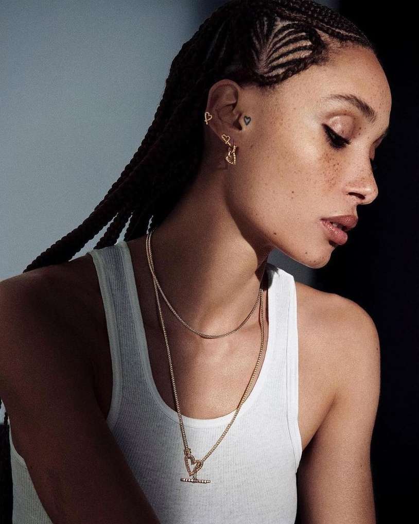 Close-up of a woman with braided hair wearing a white tank top and gold heart jewelry, showcasing beauty and fashion details.