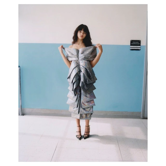 A full-length shot of an adult female posing indoors, wearing a silver gown and black high heels in front of a two-toned wall.