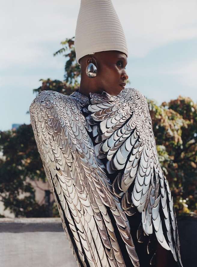 Unique fashion featuring a Black model in a silver wing-like dress and a white cone hat, posing against a blue sky and leafy trees.