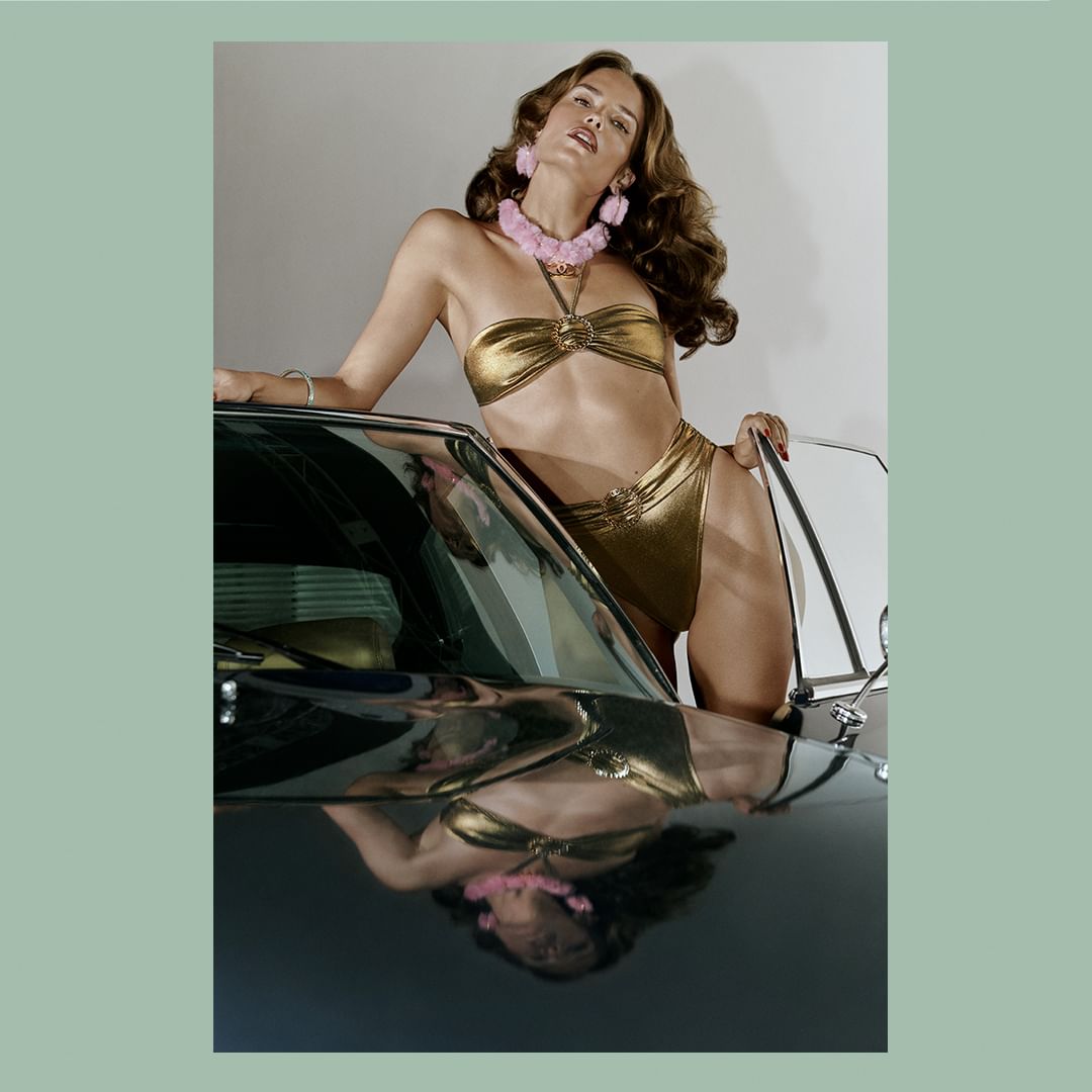 A brunette woman in a golden bikini poses on a classic car, wearing a pink fuzzy necklace and earrings, exuding a glamorous vibe.