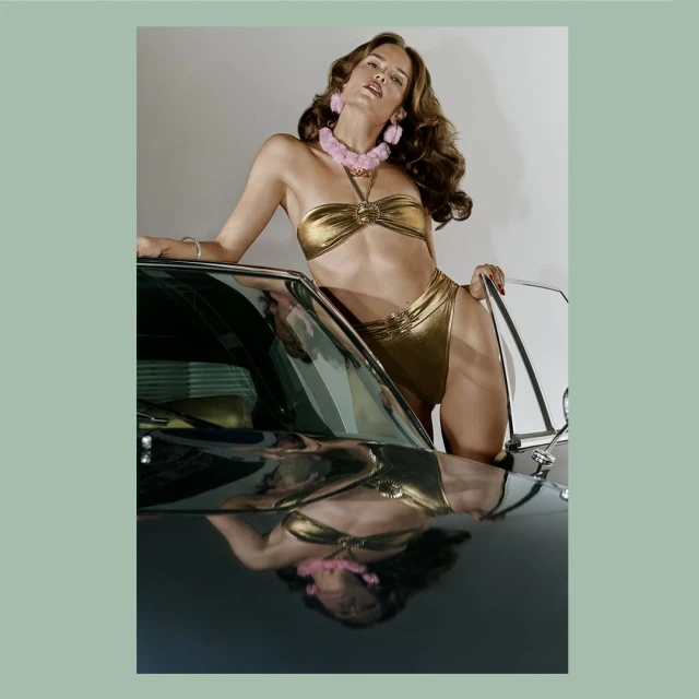 A brunette woman in a golden bikini poses on a classic car, wearing a pink fuzzy necklace and earrings, exuding a glamorous vibe.