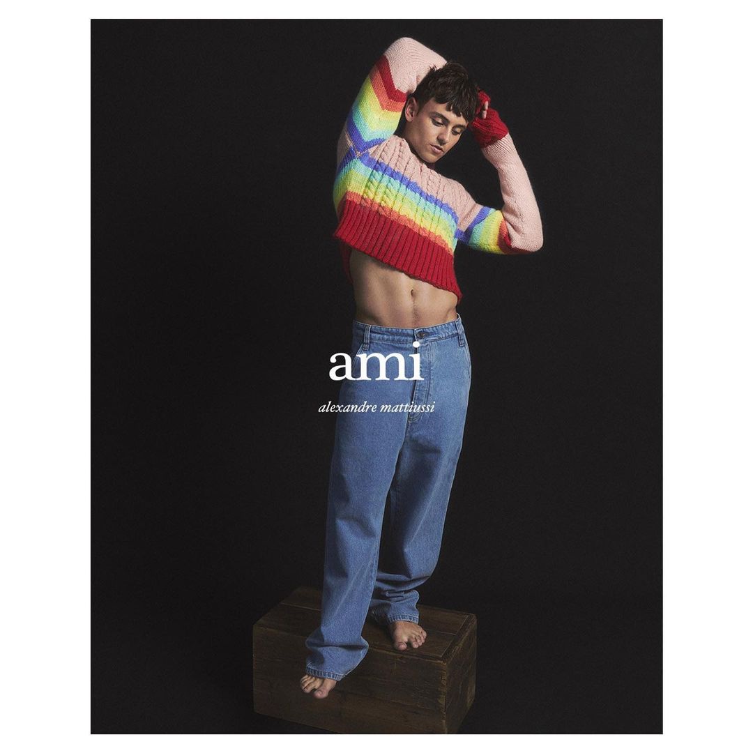 Fashion shot of a slender adult male model with an exposed torso wearing a colorful cropped sweater and jeans, standing on a wooden box.