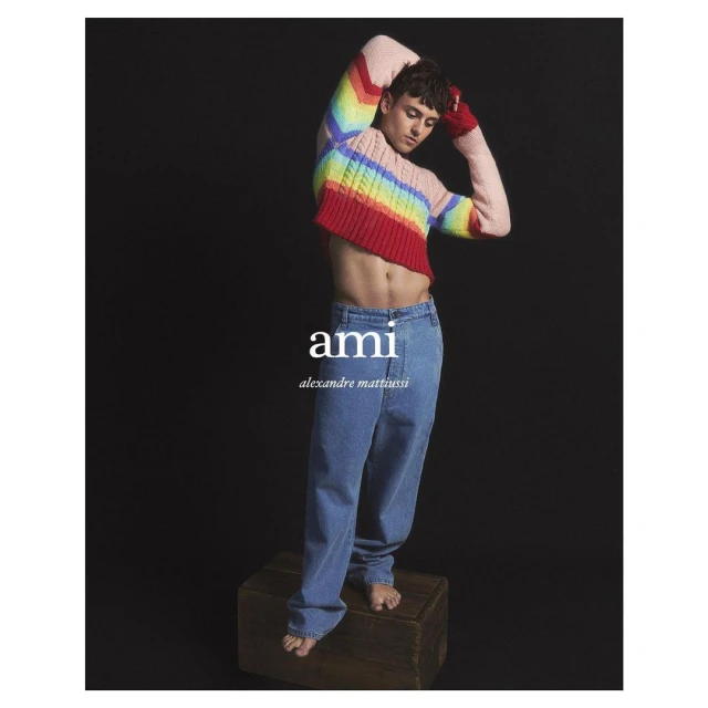 Fashion shot of a slender adult male model with an exposed torso wearing a colorful cropped sweater and jeans, standing on a wooden box.