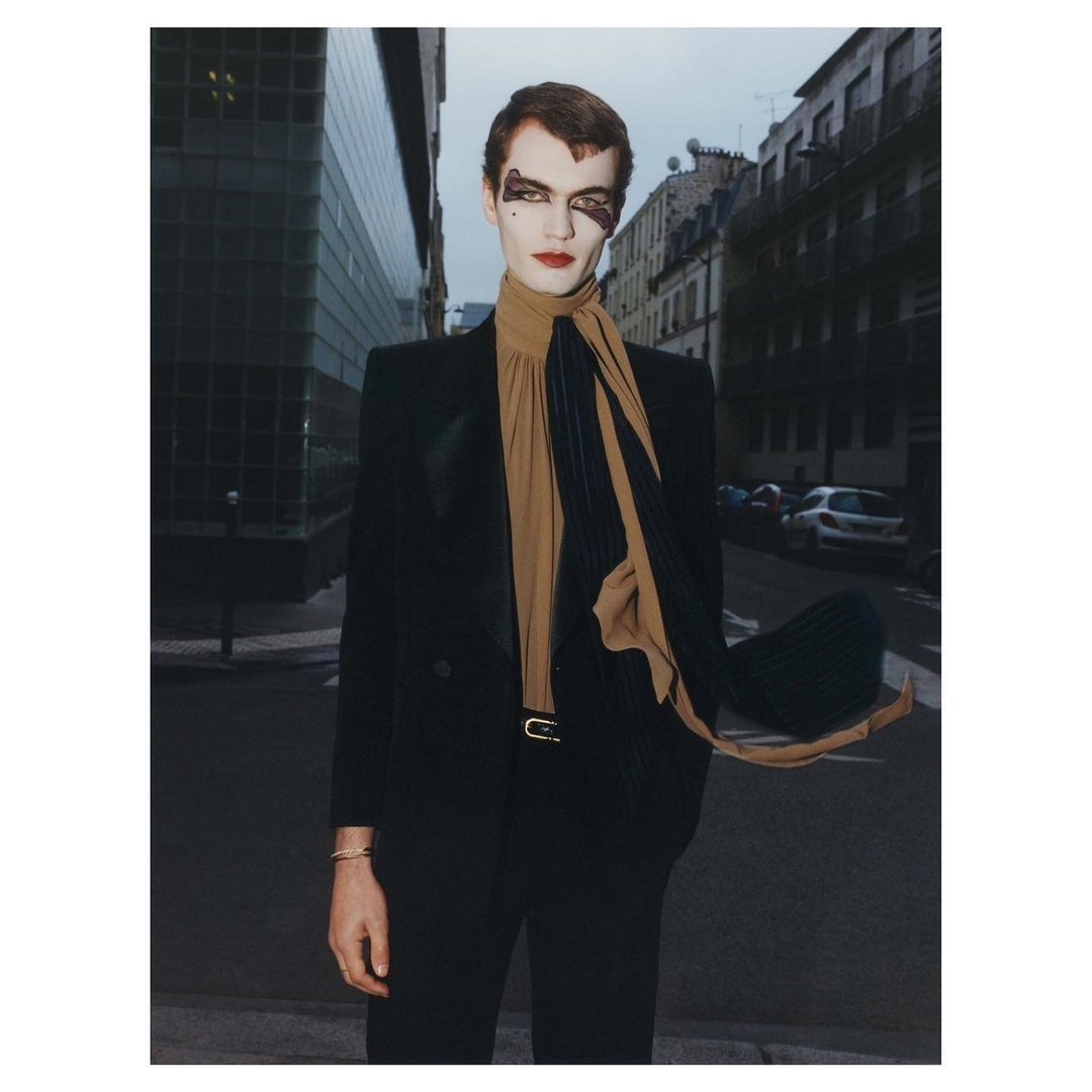 Adult male in a black suit and theatrical makeup stands on a street, wearing a beige scarf, in an urban setting on an overcast day.