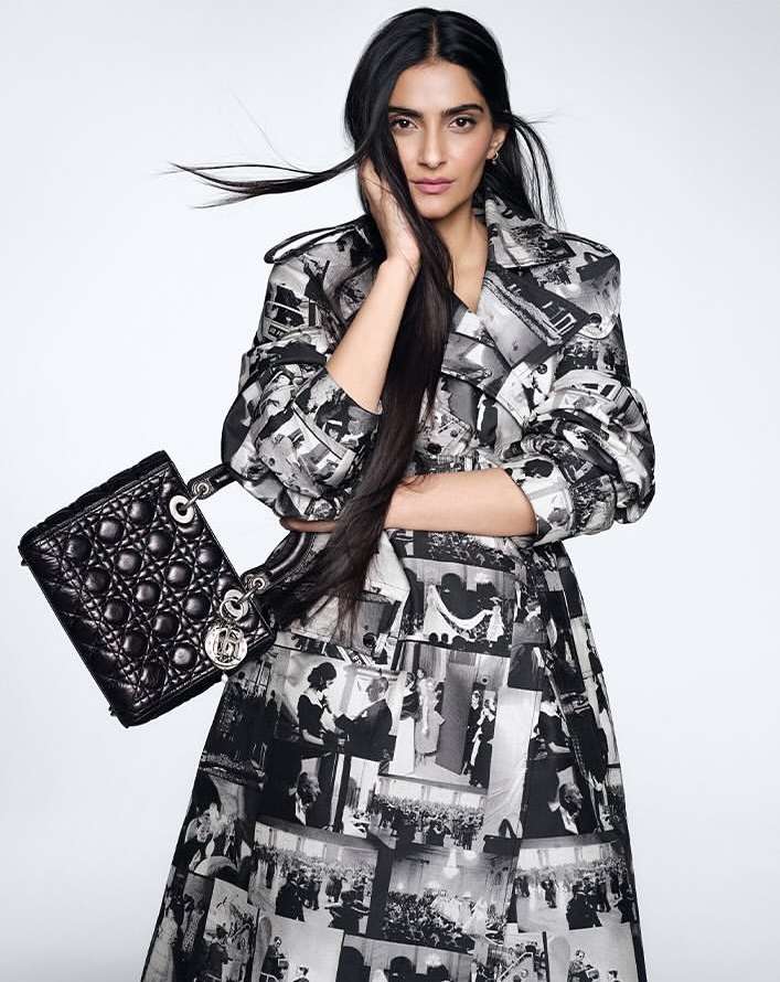 Fashionable adult female model wearing a photo-print dress and carrying a black quilted handbag in a light, neutral studio.