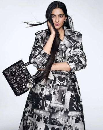 Fashionable adult female model wearing a photo-print dress and carrying a black quilted handbag in a light, neutral studio.