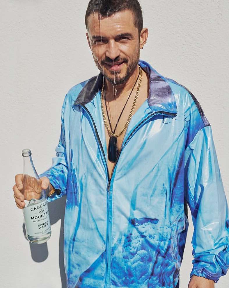 Man in blue patterned jacket, water pouring on head, holding water bottle against a white background. He has a gold chain.