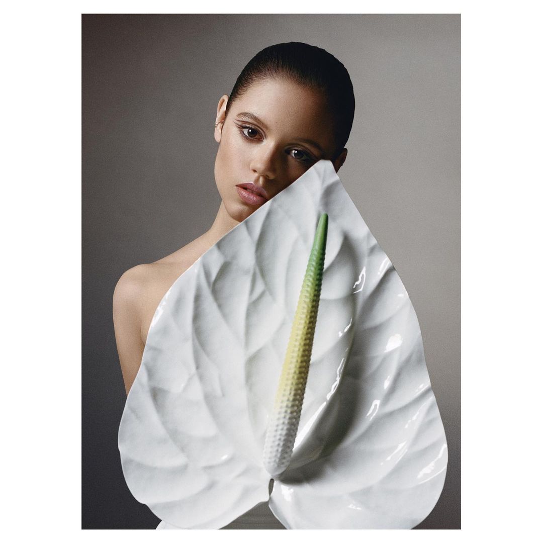 Close-up portrait of a young woman posing with a large white Anthurium flower against a soft grey background in a beauty shot.