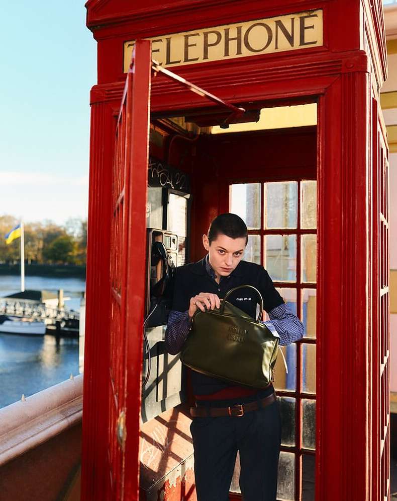 Young adult stands in a red telephone booth with a bag, city and river views in background under a bright sky. Fashion shot.