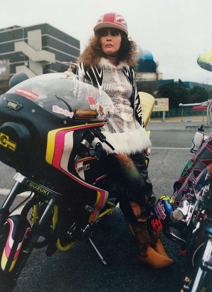 Woman leaning on motorcycle with retro styling, wearing a Union Jack hat, shiny jacket, and cowboy boots in an outdoor setting.
