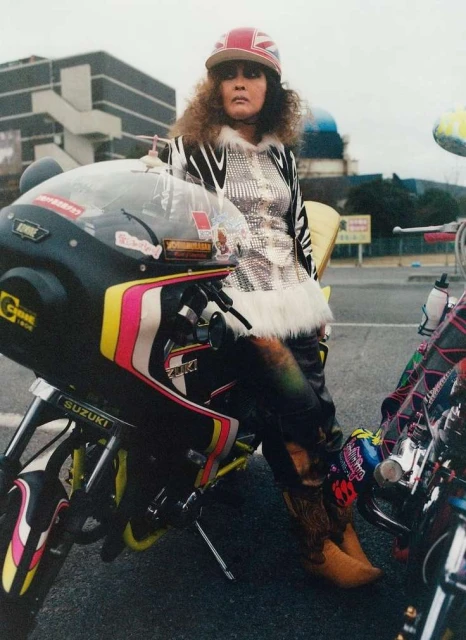 Woman leaning on motorcycle with retro styling, wearing a Union Jack hat, shiny jacket, and cowboy boots in an outdoor setting.