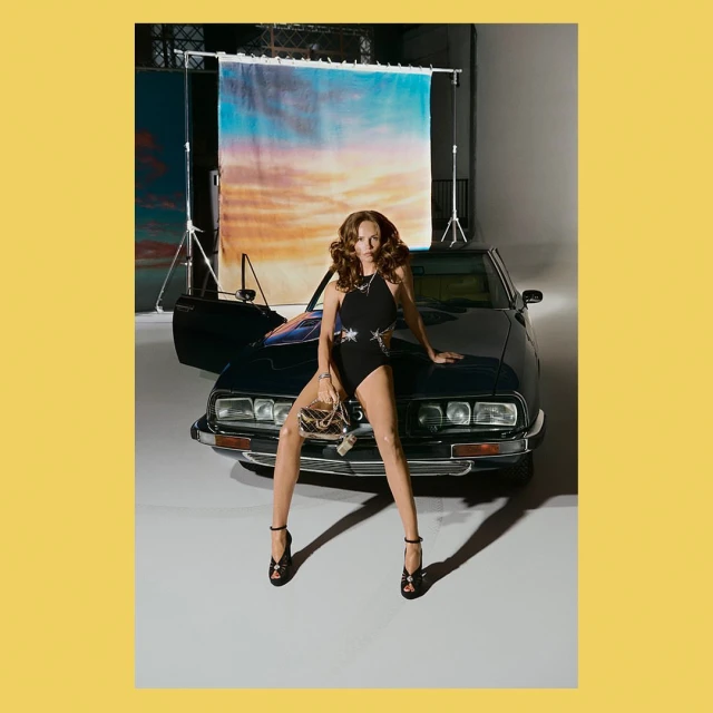 Glamorous adult female model poses on a car in a black leotard and heels, holding a purse with a backdrop in a photo studio.
