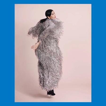 Woman in a shiny silver fringed coat jumps in a pink studio. High fashion, studio shot, light pink backdrop, full body, fashion concept.