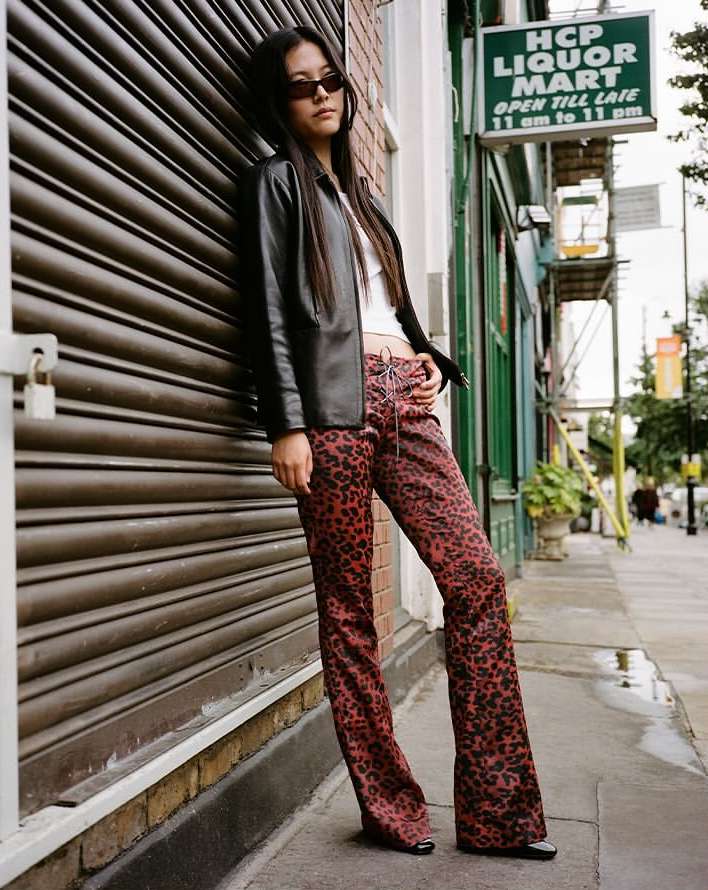 Woman in sunglasses poses in front of roll-up door in a black jacket and red leopard print pants, standing on city sidewalk.