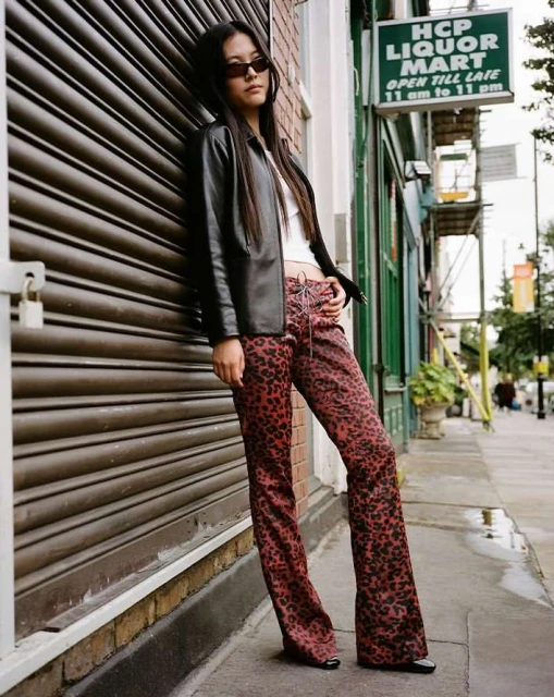 Woman in sunglasses poses in front of roll-up door in a black jacket and red leopard print pants, standing on city sidewalk.